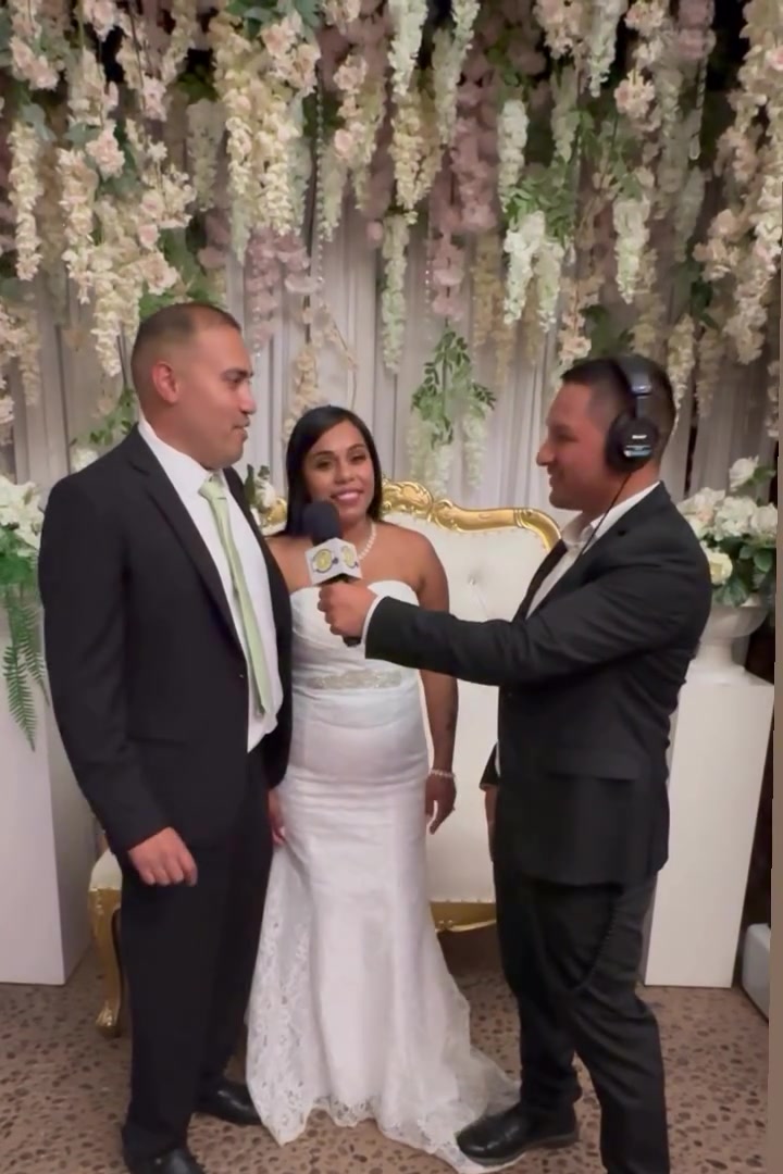 The DJ on the mic with a bride and groom in front of a floral wall